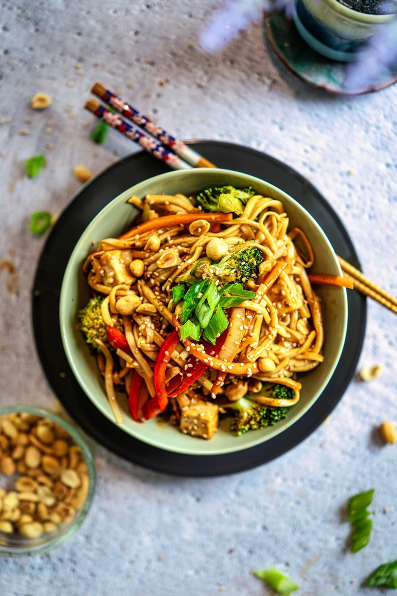 A bowl of udon noodles mixed with vegetables and topped with peanuts and herbs, accompanied by chopsticks.