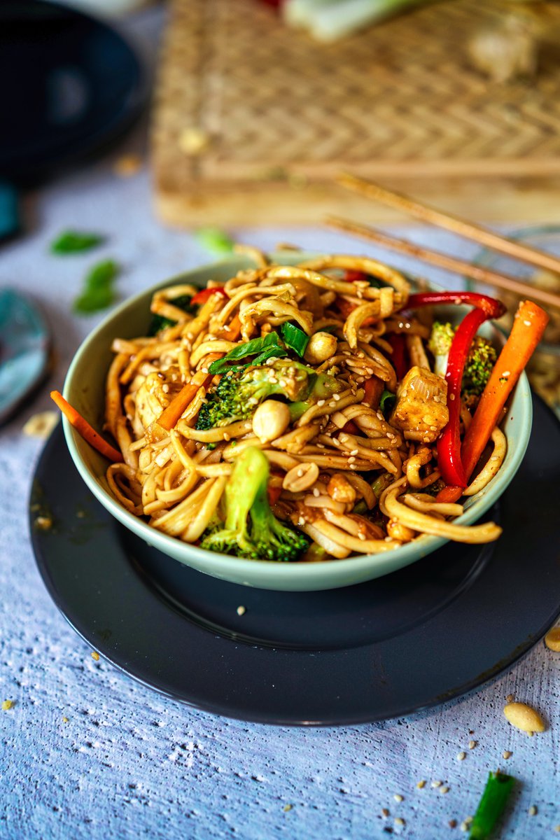 Bowl of udon noodles stir-fried with vegetables and peanuts, garnished with sesame seeds.