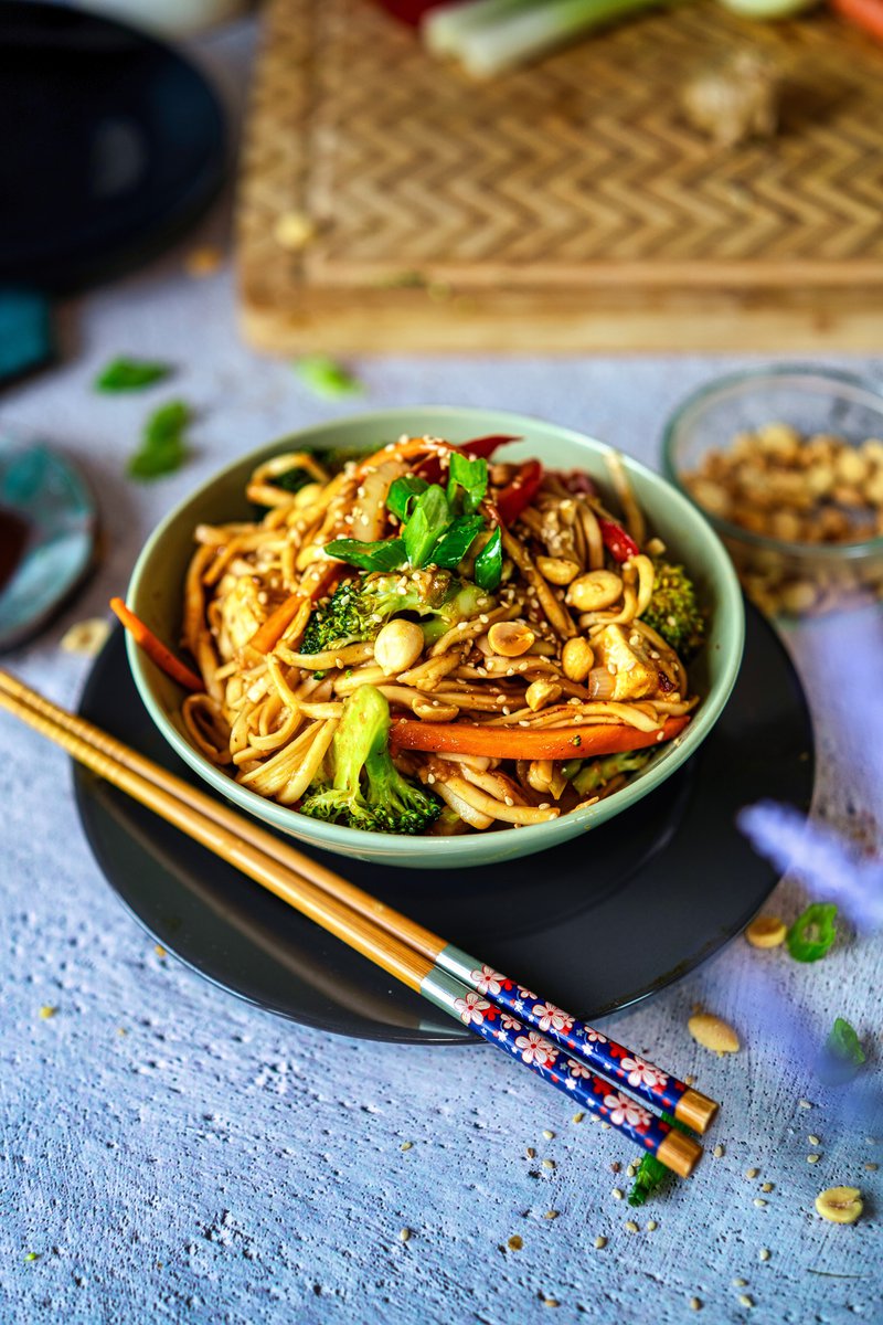 A bowl of udon noodles with vegetables and peanuts, garnished with herbs, accompanied by a pair of decorated chopsticks.