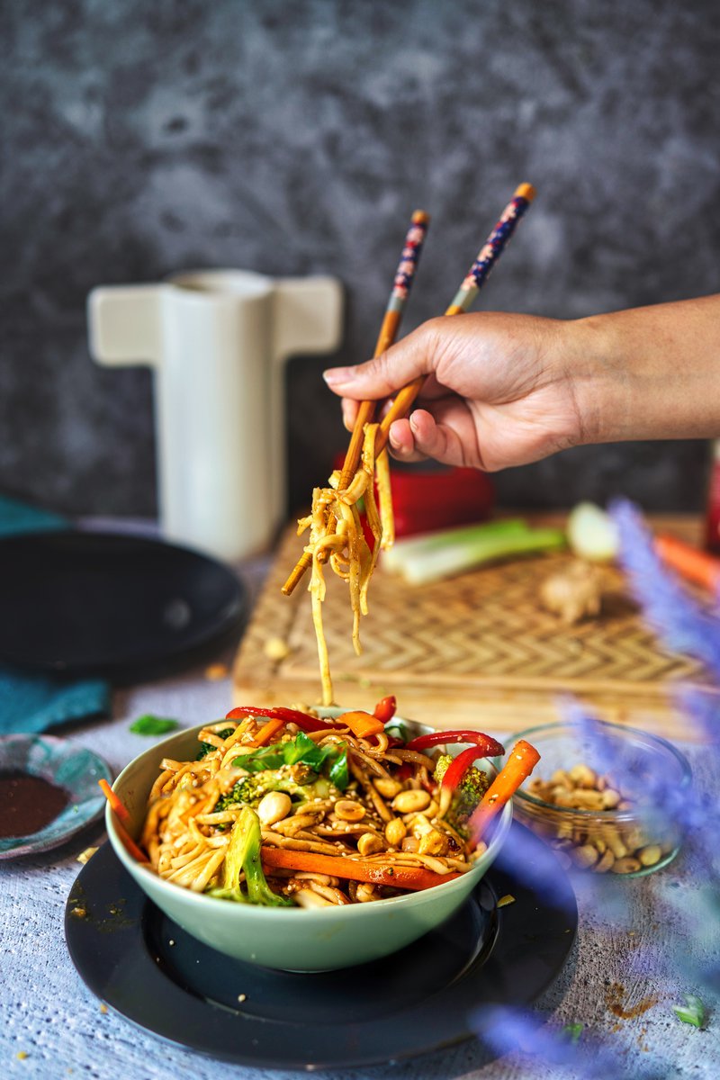 A hand using chopsticks to lift udon noodles from a bowl filled with colorful vegetables and garnished with peanuts.