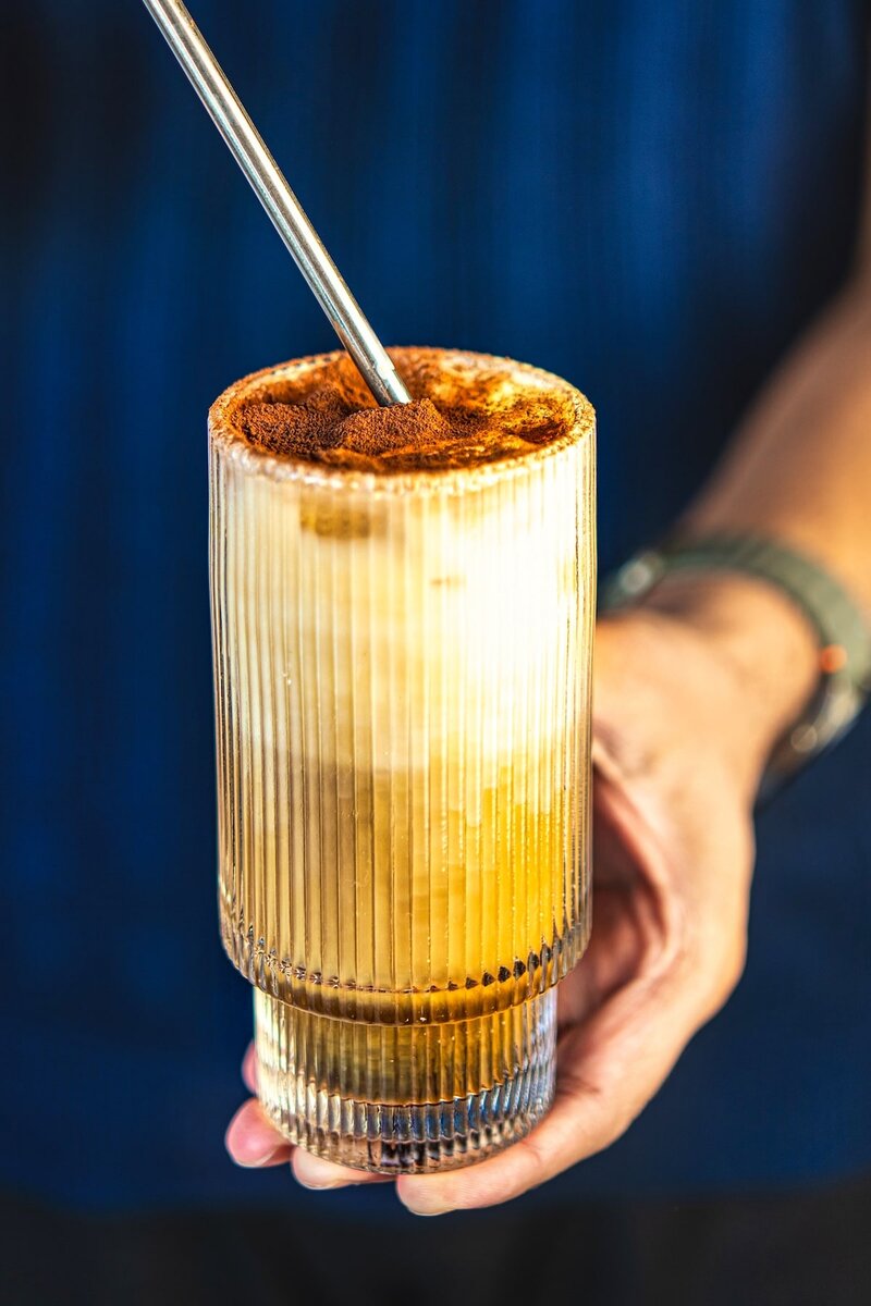 Hand holding a creamy vanilla iced latte with foam and cocoa dust, refreshing iced coffee drink close-up