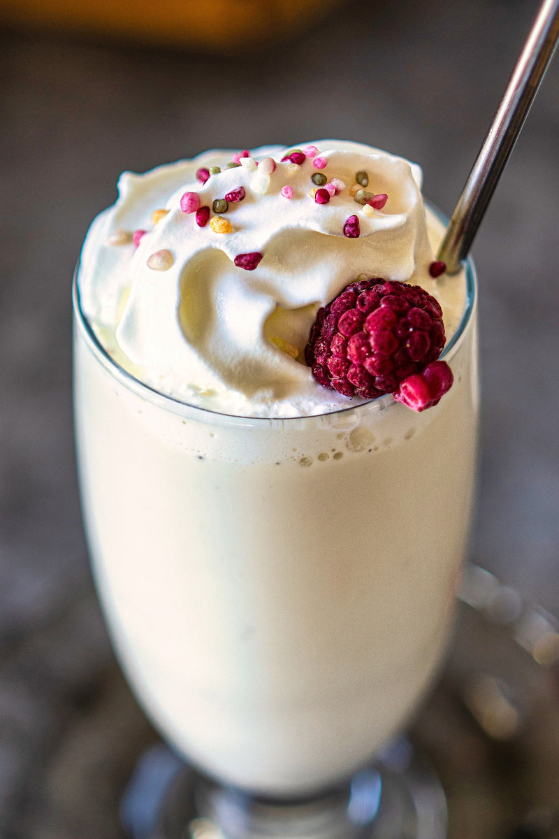 A close-up image of a vanilla milkshake in a tall glass, topped with whipped cream, a frozen raspberry, and colorful sprinkles. The drink is served with a straw.