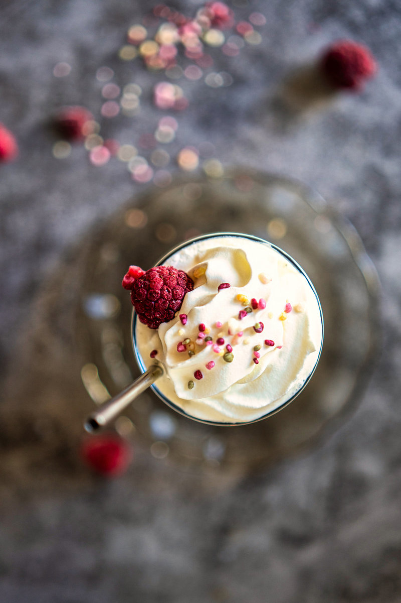 Top-down view of a vanilla milkshake in a glass, garnished with whipped cream, a frozen raspberry, and multicolored sprinkles, seen from above on a grey textured background with blurred raspberries and sparkling decorations scattered around