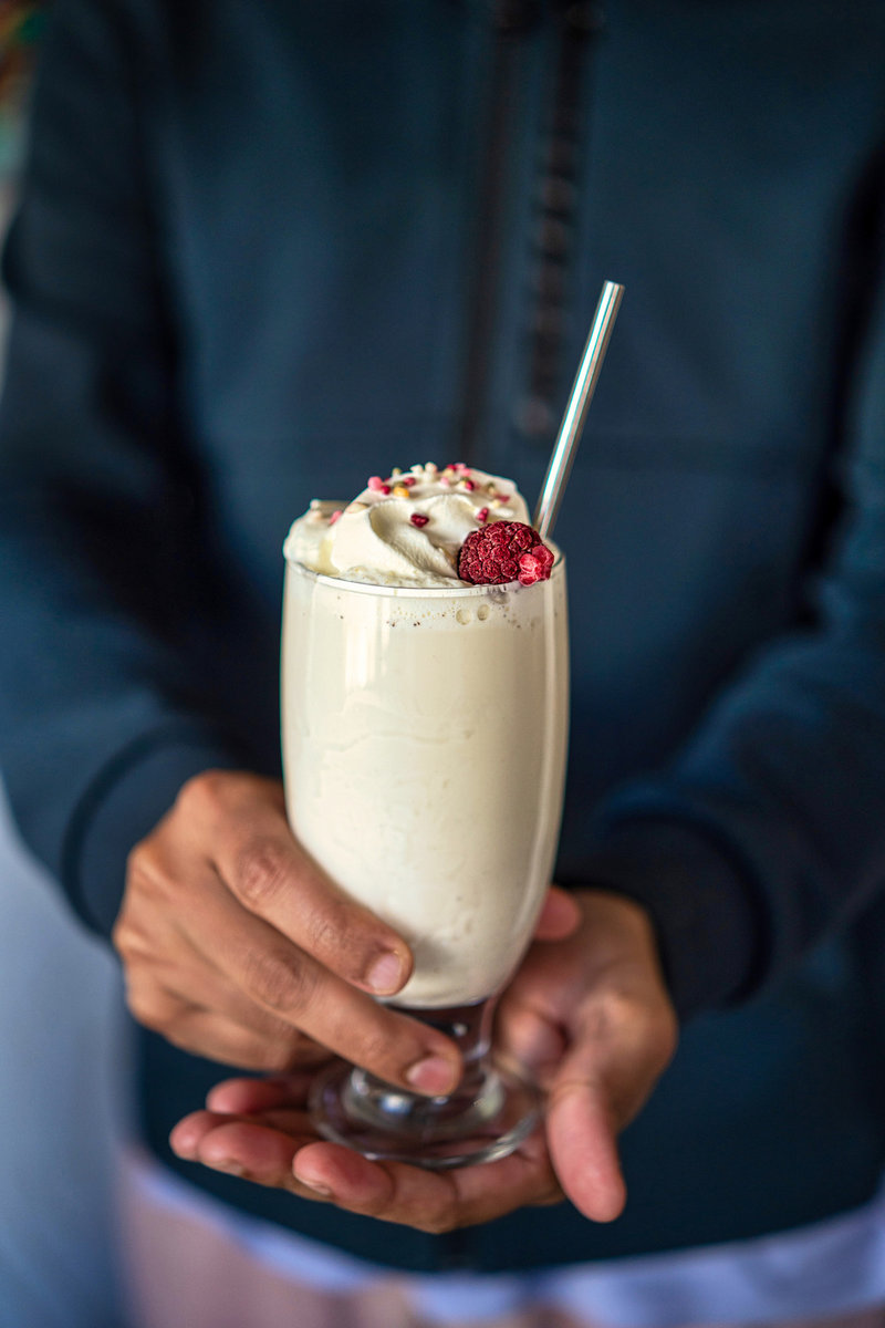 Man in a dark blue jacket holding a vanilla milkshake garnished with whipped cream, frozen raspberries, and colorful sprinkles, served in a tall glass with a metal straw.