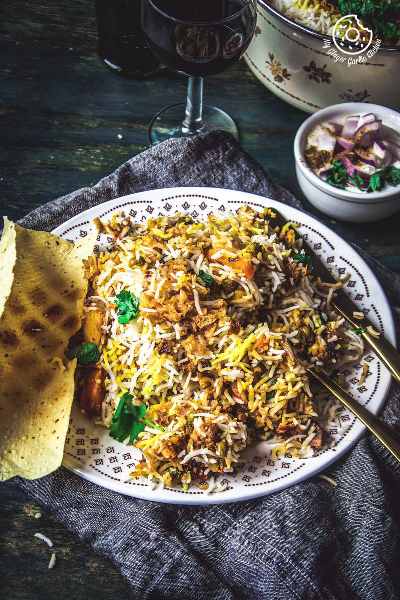 there is a plate of restaurant style hyderabadi veg dum biryani with a papad and a glass of wine