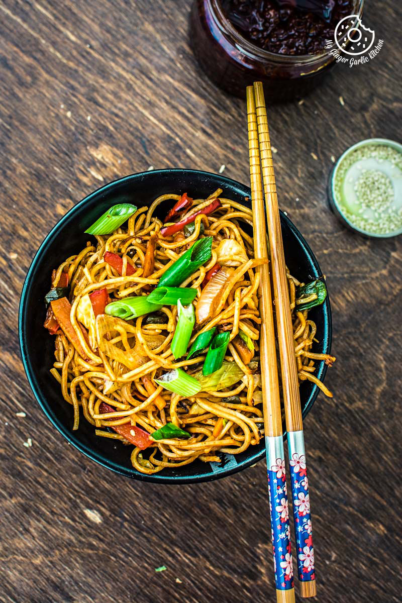 a close up of a bowl of veg schezwan noodles with chopsticks on a table