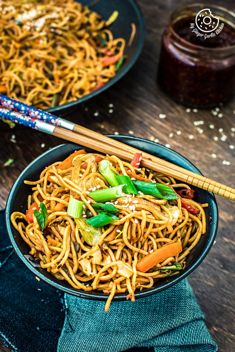 a close up of a bowl of veg schezwan noodles with spring onions and chopsticks on a table