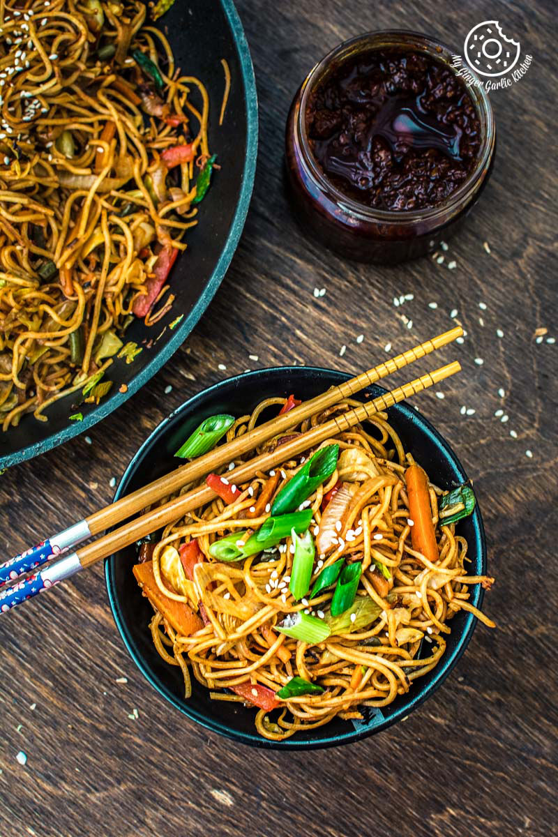 a close up of a bowl of veg schezwan noodles with spring onions, chopsticks and a bowl of sauce