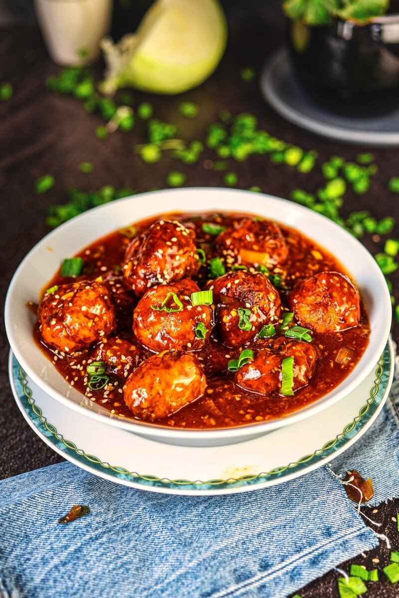 Veg manchurian gravy served in a bowl, crispy vegetable balls coated in a glossy spicy garlic sauce with spring onions and sesame seeds