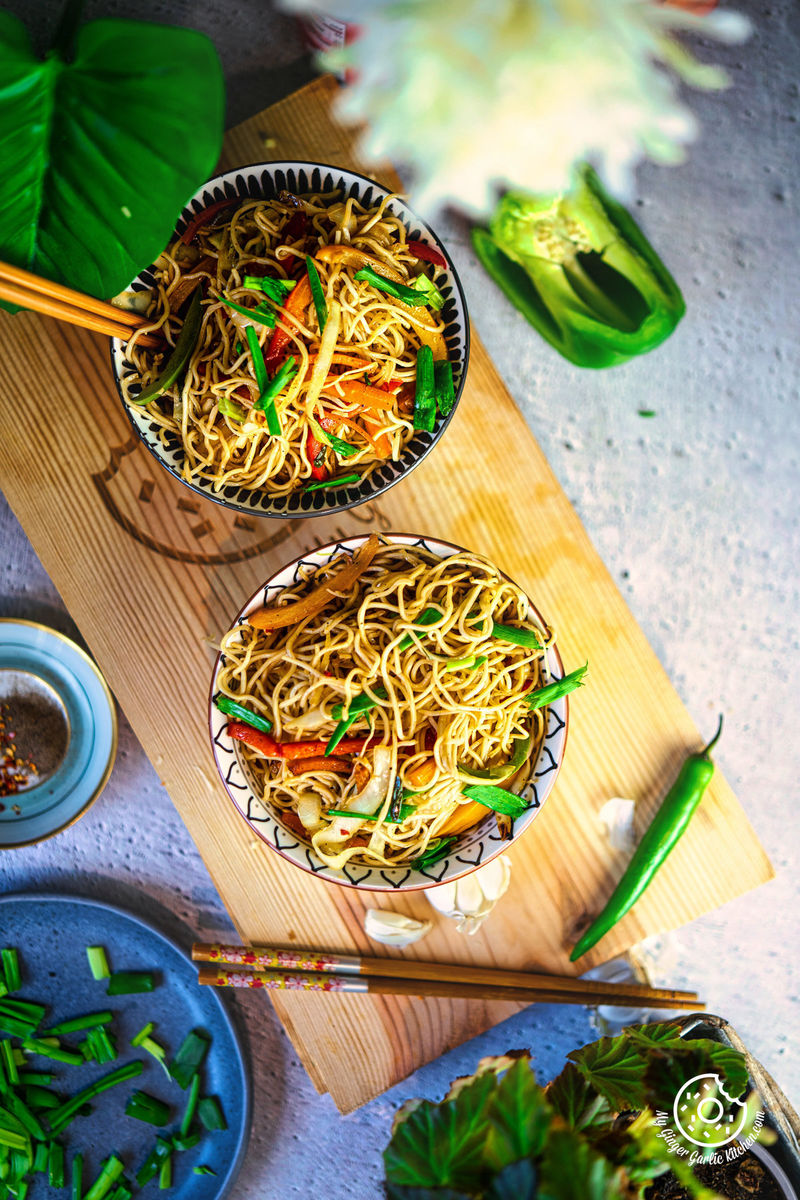 overhead view of two bowls of vegetable hakka noodles on a wooden board