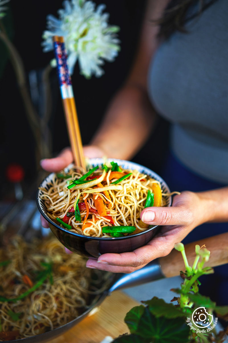 a person holding a black bowl with veg hakka noodles