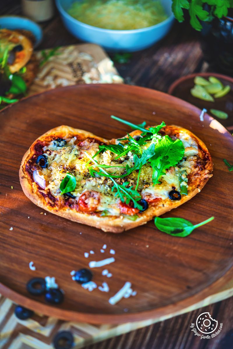 1 vegetarian heart pizza topped with green leaves sitting on top of a wooden plate with olives on the side