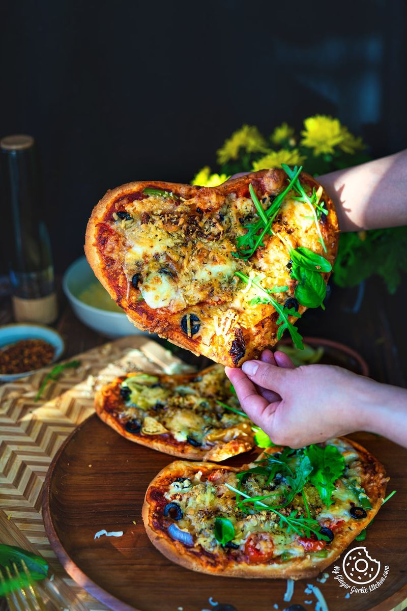 a person holding 1 vegetarian heart pizza topped with green leaves over 2 vegetarian heart pizzas on a wooden plate