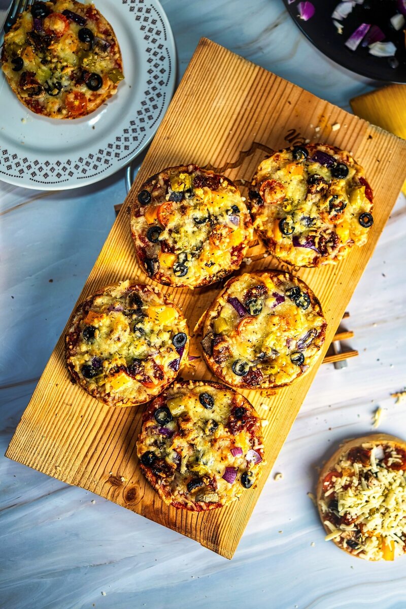 Homemade veggie bum pizzas with cheese, olives and vegetables on rustic wooden board next to decorative plate, perfect for easy healthy appetizers