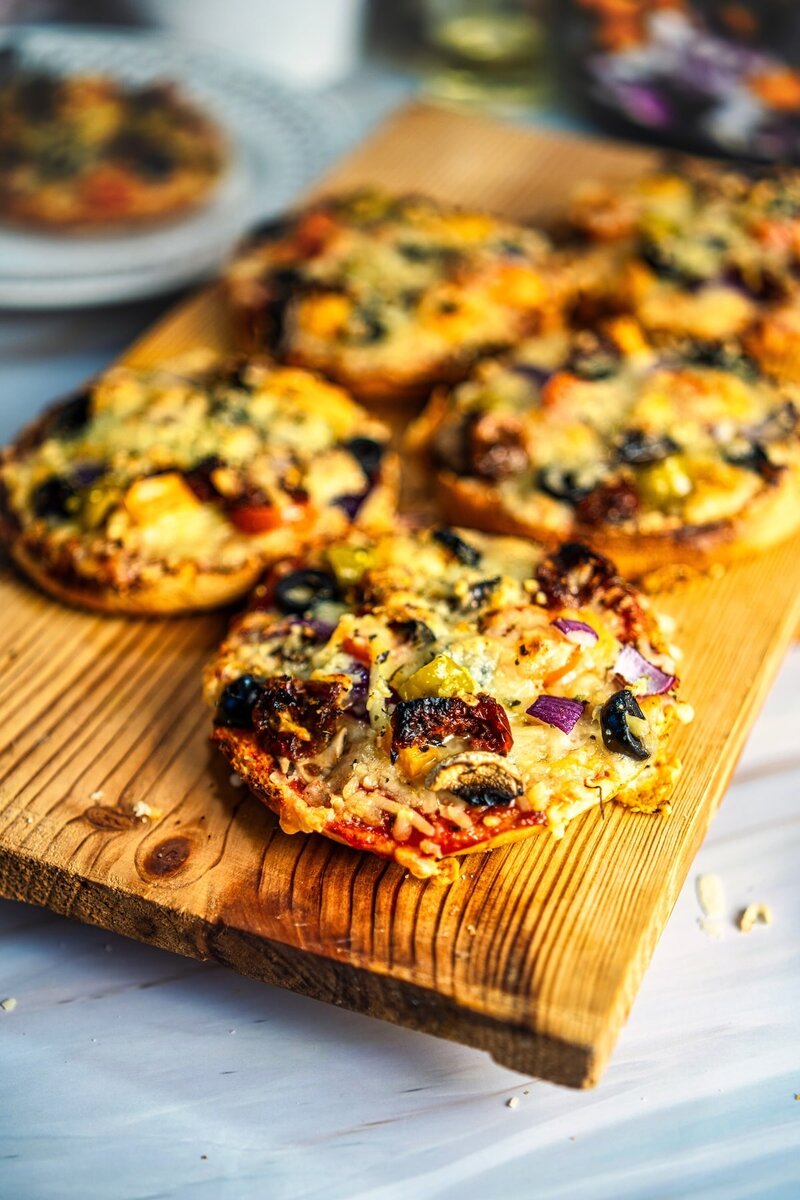Close up of crispy mini veggie bun pizzas topped with cheese, olives, and vegetables on a rustic wooden serving board