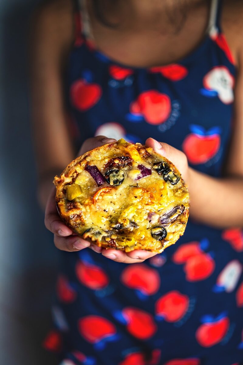 Child holding a freshly baked veggi bun pizza with cheese, black olives, and vegetables, kid friendly homemade snack