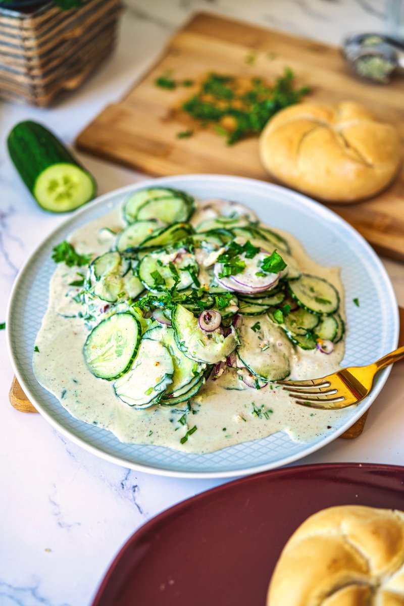 A creamy cucumber salad with red onion and fresh herbs served on a plate, next to a sliced cucumber and a bread roll in the background.