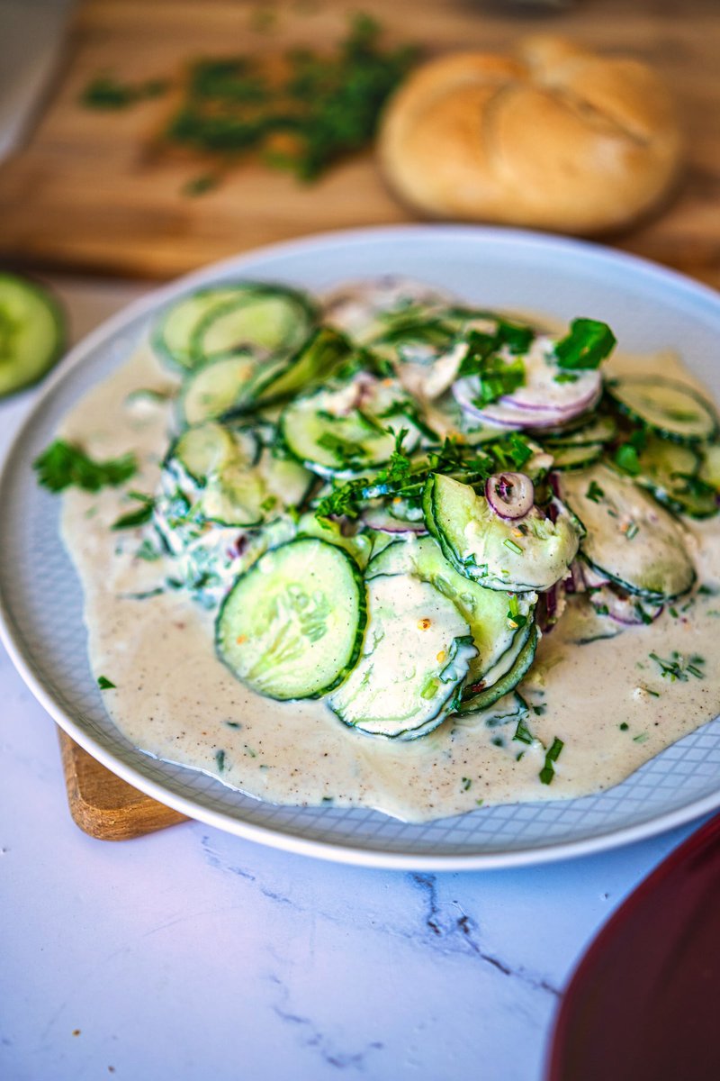 Close-up of cucumber slices and red onion in a creamy yogurt-based dressing, garnished with cilantro, served on a plate with a bread roll in the background.