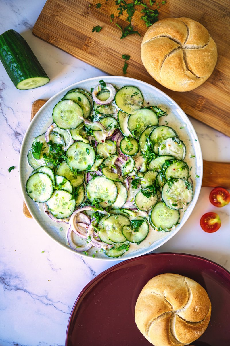  Creamy cucumber salad in a white bowl, surrounded by fresh ingredients and kaiser rolls on a marble surface.