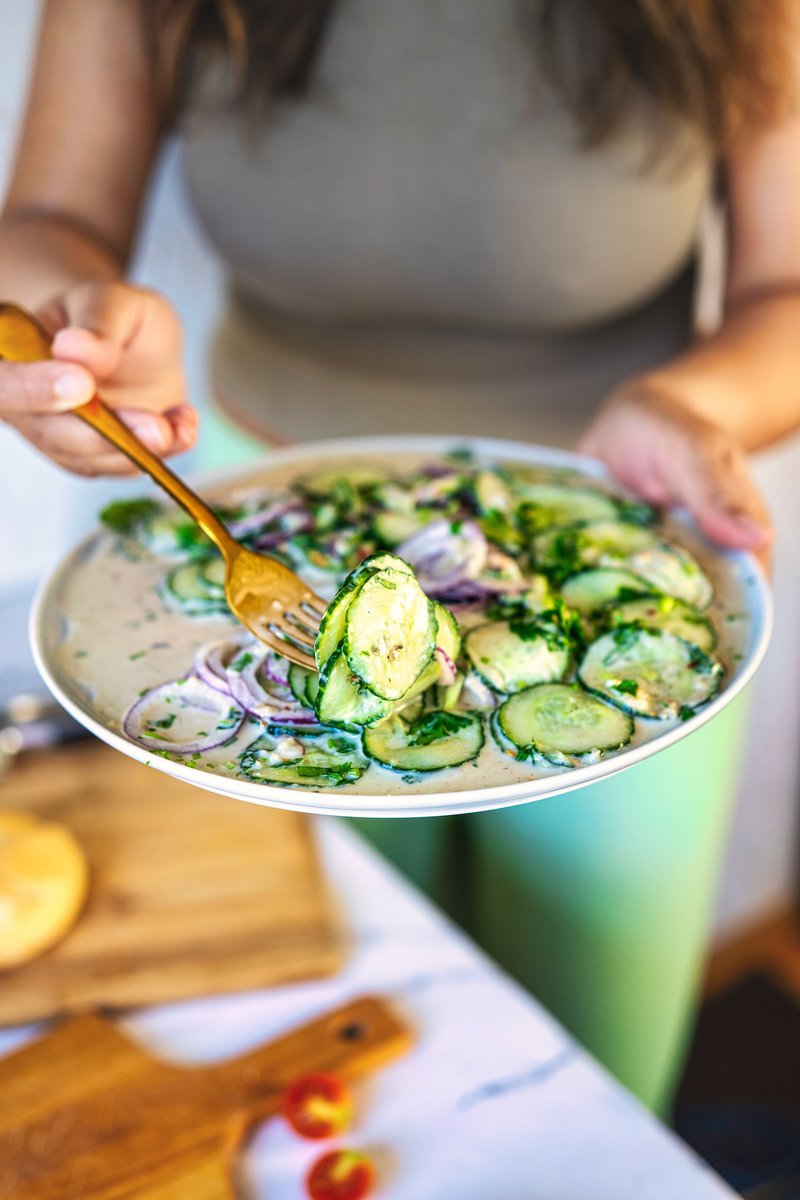 A person holding a fork with a slice of cucumber from a creamy cucumber salad with red onion and herbs on a plate, with a wooden cutting board and tomatoes in the background.