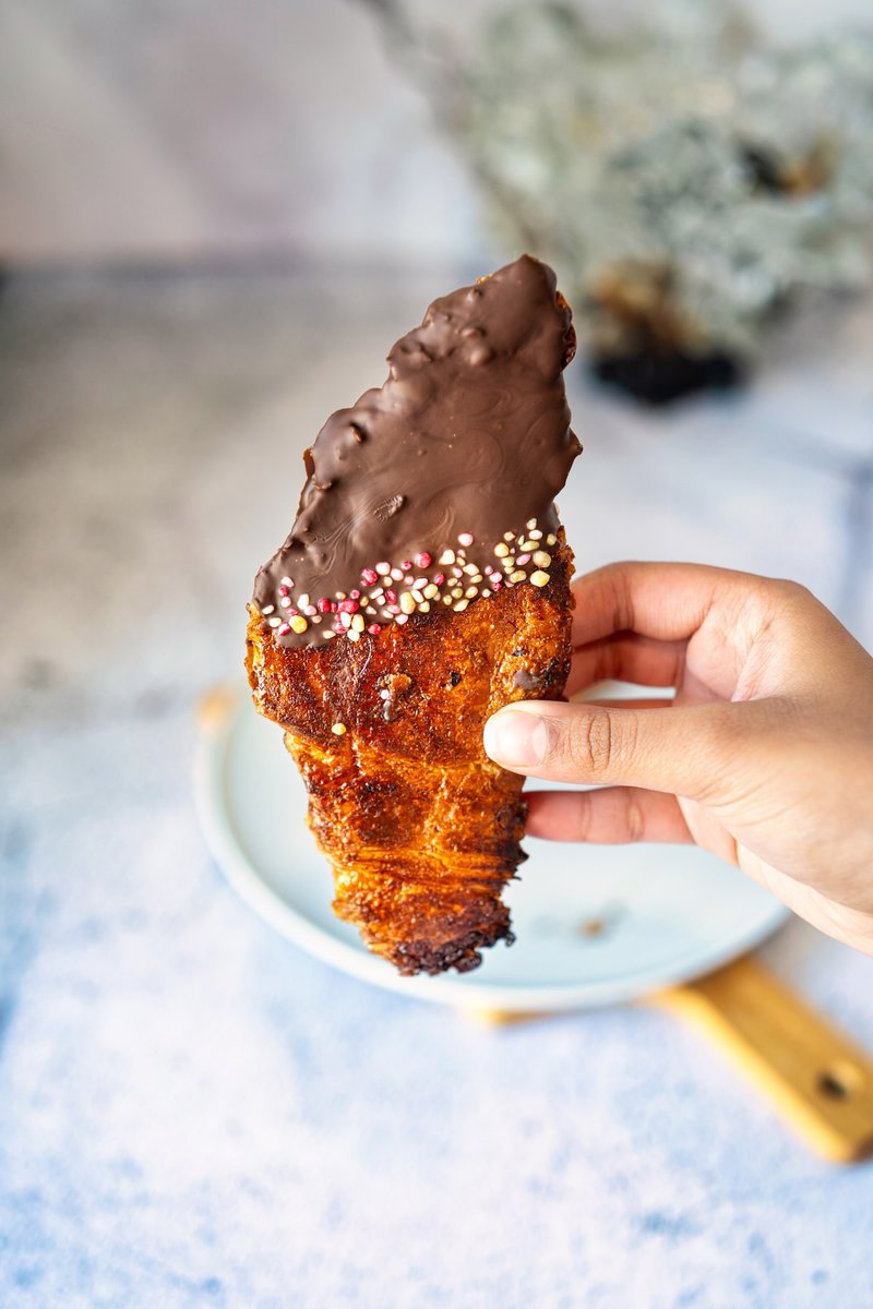A hand holding a flat croissant half-dipped in chocolate and decorated with multicolored sprinkles.