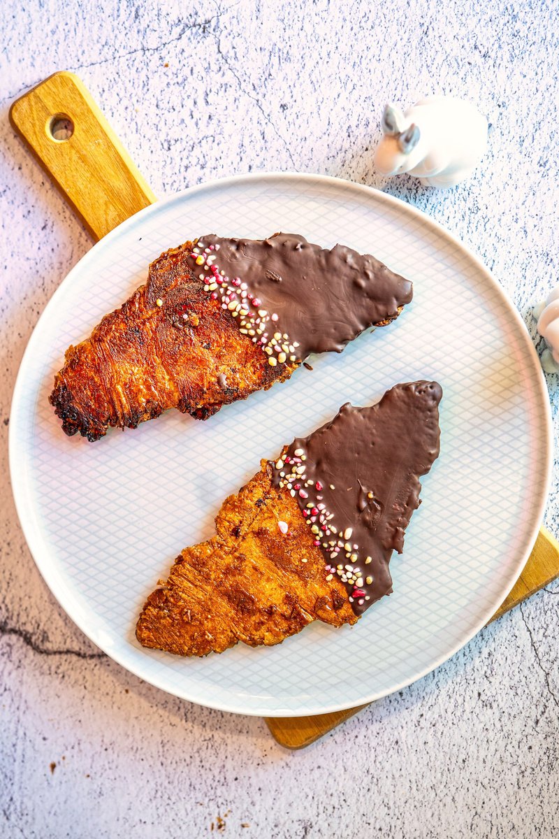 Two flat croissants covered in chocolate and colorful sprinkles displayed on a round plate with a wooden cutting board.