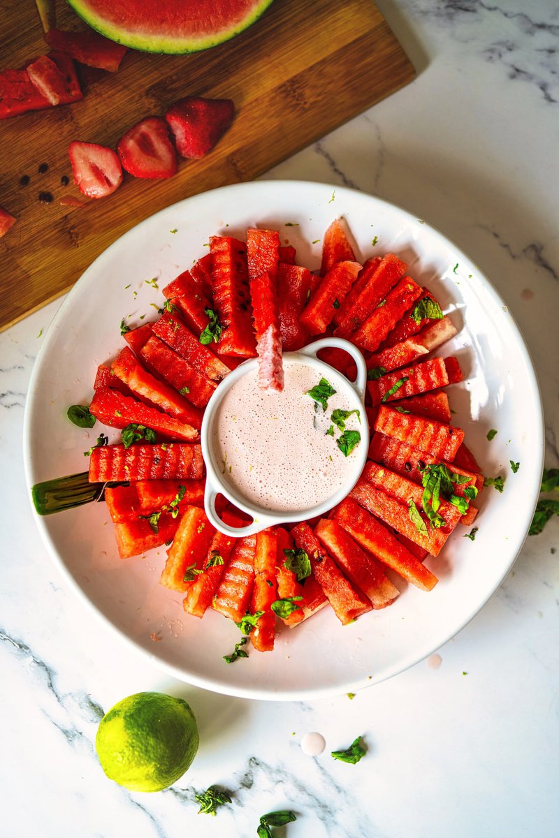 A close-up of watermelon fries arranged around a bowl of strawberry yogurt dip, garnished with mint and lime zest.