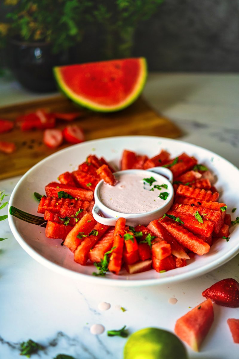 IA side view of watermelon fries with strawberry yogurt dip, with sliced strawberries and watermelon in the background.