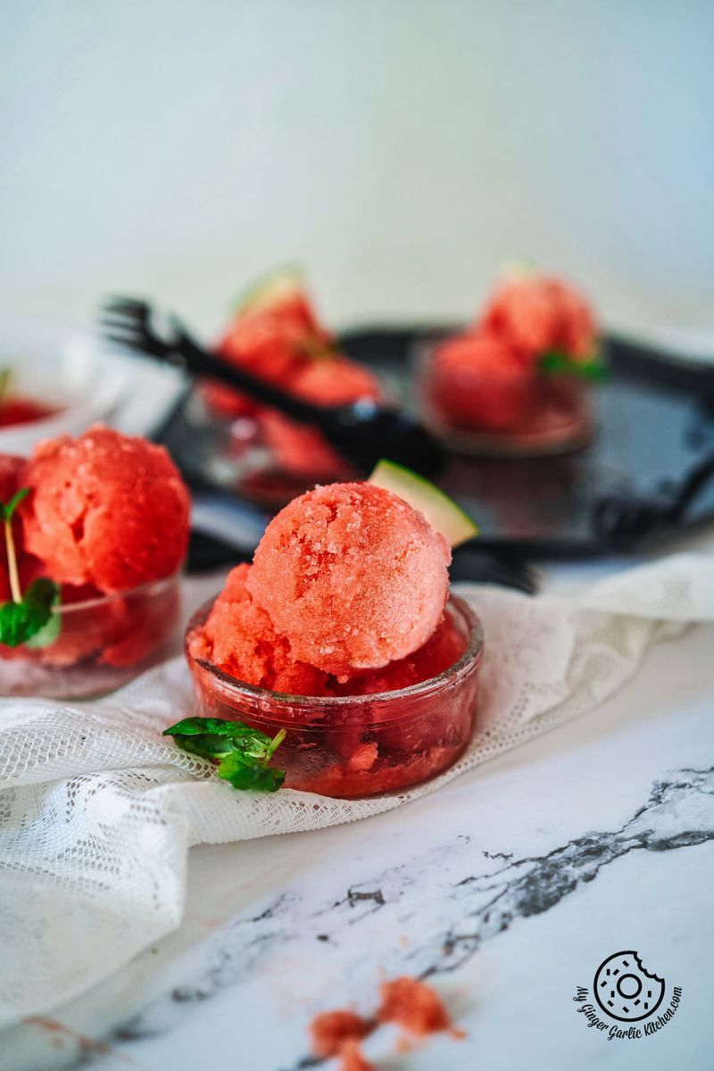 2 watermelon sorbet scoops in a transparent bowl and 3 bowls in the background
