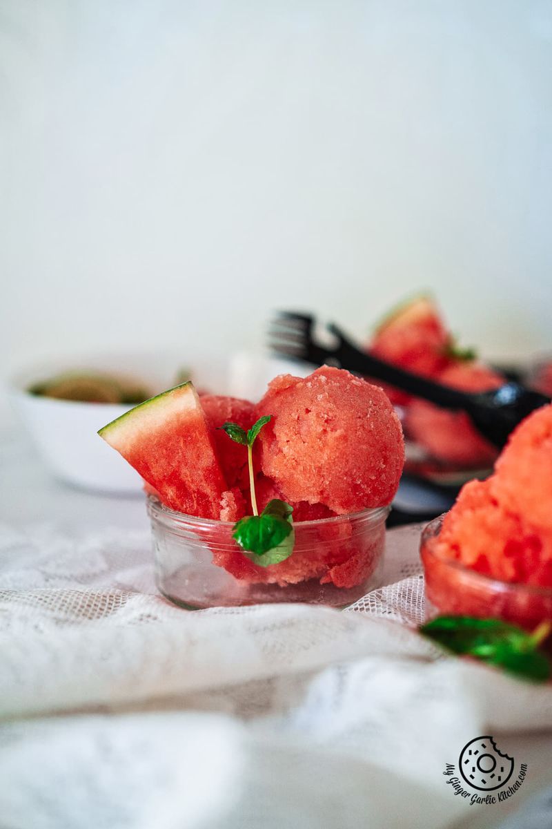 2 watermelon sorbet scoops in a tarnsparent bowl with watermelon slice and mint sprig