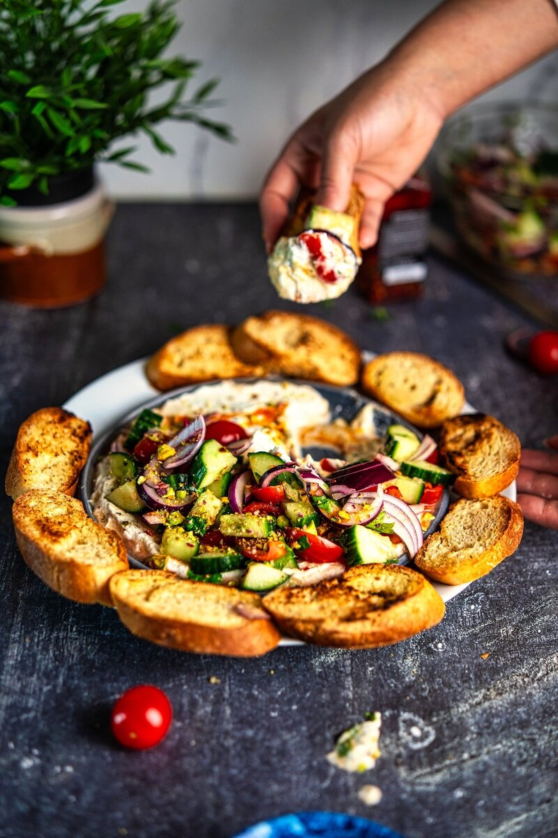Hand holding toasted baguette topped with whipped cottage cheese dip and fresh tomato cucumber salad