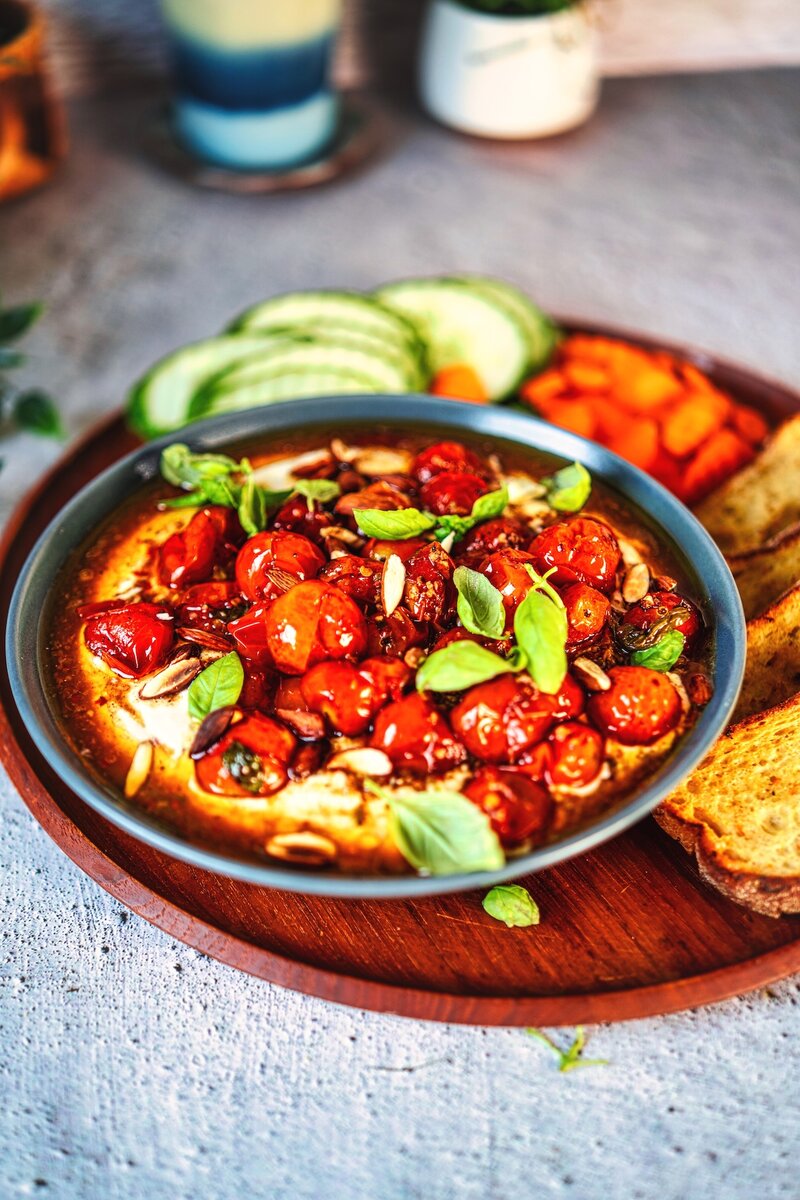 Mediterranean whipped feta cheese dip topped with caramelized cherry tomatoes, sliced almonds, and basil leaves on wooden serving board