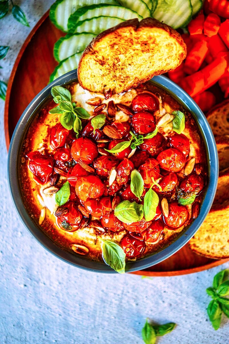 Whipped feta dip with roasted cherry tomatoes, almonds, and fresh basil served in black bowl with toasted bread and cucumber slices