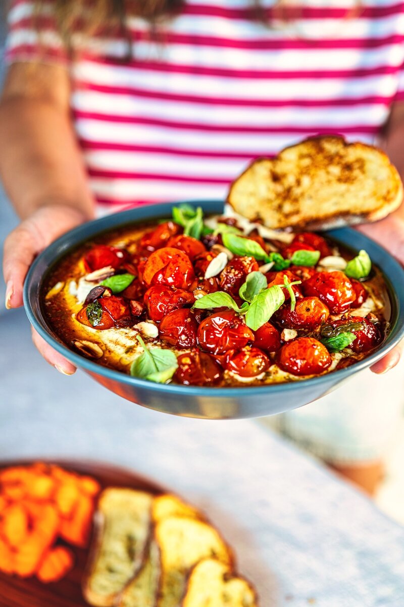 Person holding bowl of creamy whipped feta dip with roasted cherry tomatoes, nuts, and basil garnish - easy Mediterranean appetizer recipe