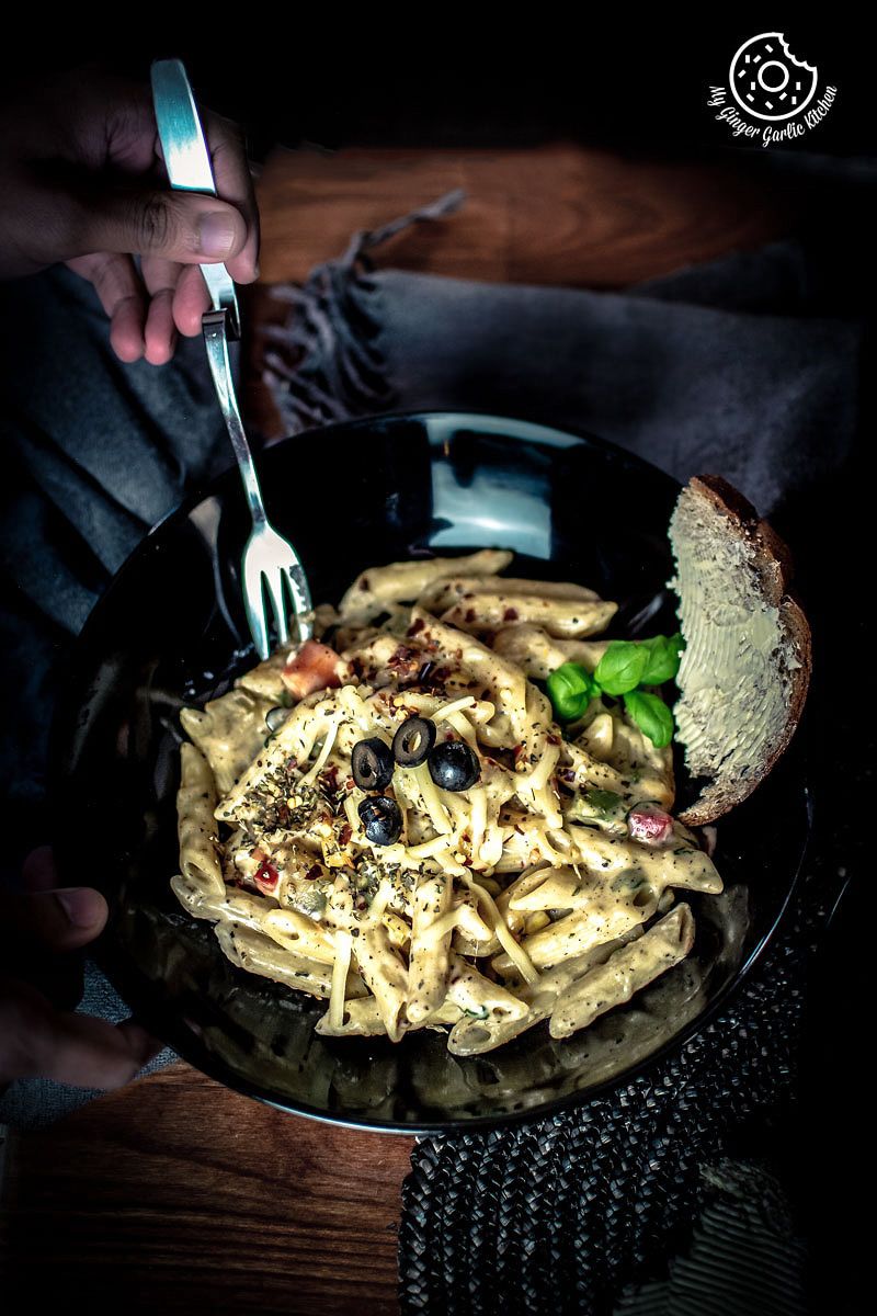 someone holding a fork over white sauce pasta bowl with black olives