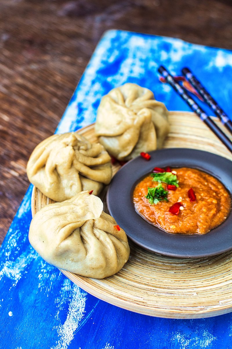 A plate of whole wheat steamed dumplings (momos) served with spicy tomato chutney, with chopsticks on the side, on a rustic wooden table with a blue napkin under the plate.