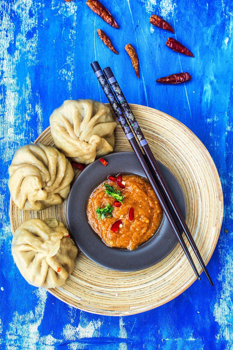 A vibrant presentation of whole wheat momos on a circular bamboo plate, with a bowl of red chutney in the center, surrounded by scattered dried red chilies on a blue wooden background.