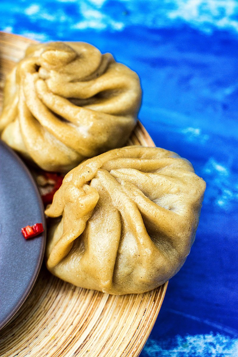Close-up of two whole wheat momos on a bamboo plate, highlighting the intricate folds of the dough, with a splash of red chutney visible in the background, all against a bright blue painted surface.