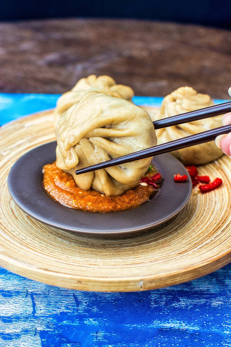 A hand using black chopsticks to pick up a wheat momo above a bowl of spicy red chutney on a bamboo plate, with a vivid blue wooden backdrop.
